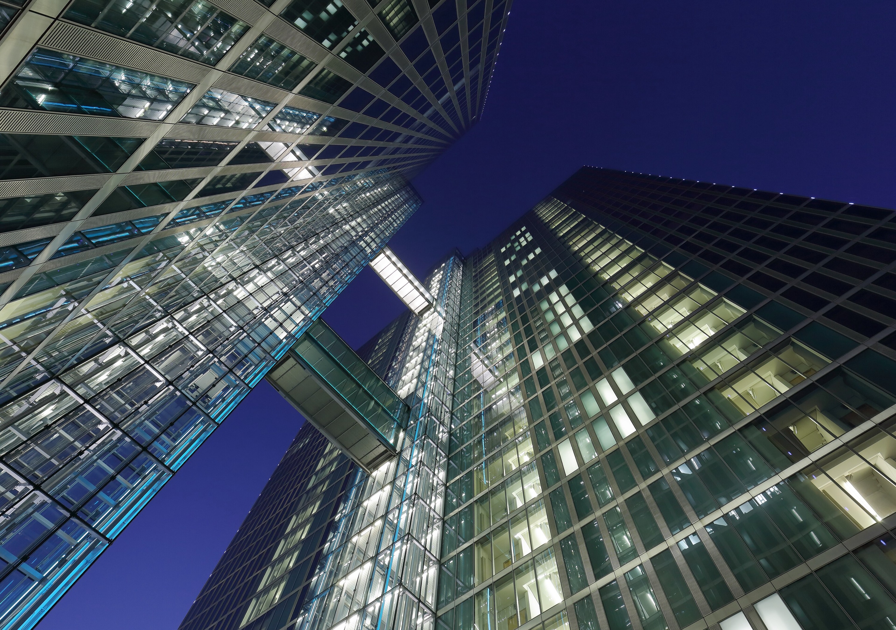 A modern glass tower at dusk, windows lit, seen from below